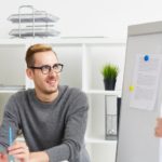 Man at a desk discussing a project with a colleague beside a whiteboard